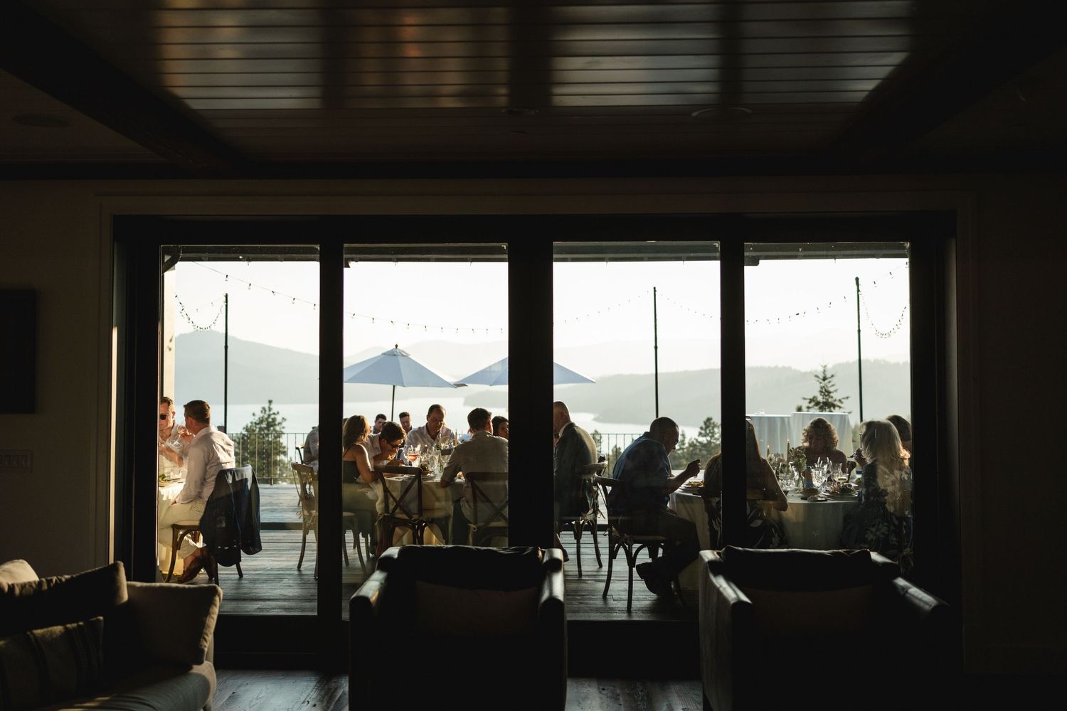 Silhouette of guests dining on an outdoor terrace with stunning mountain lake views through glass doors.