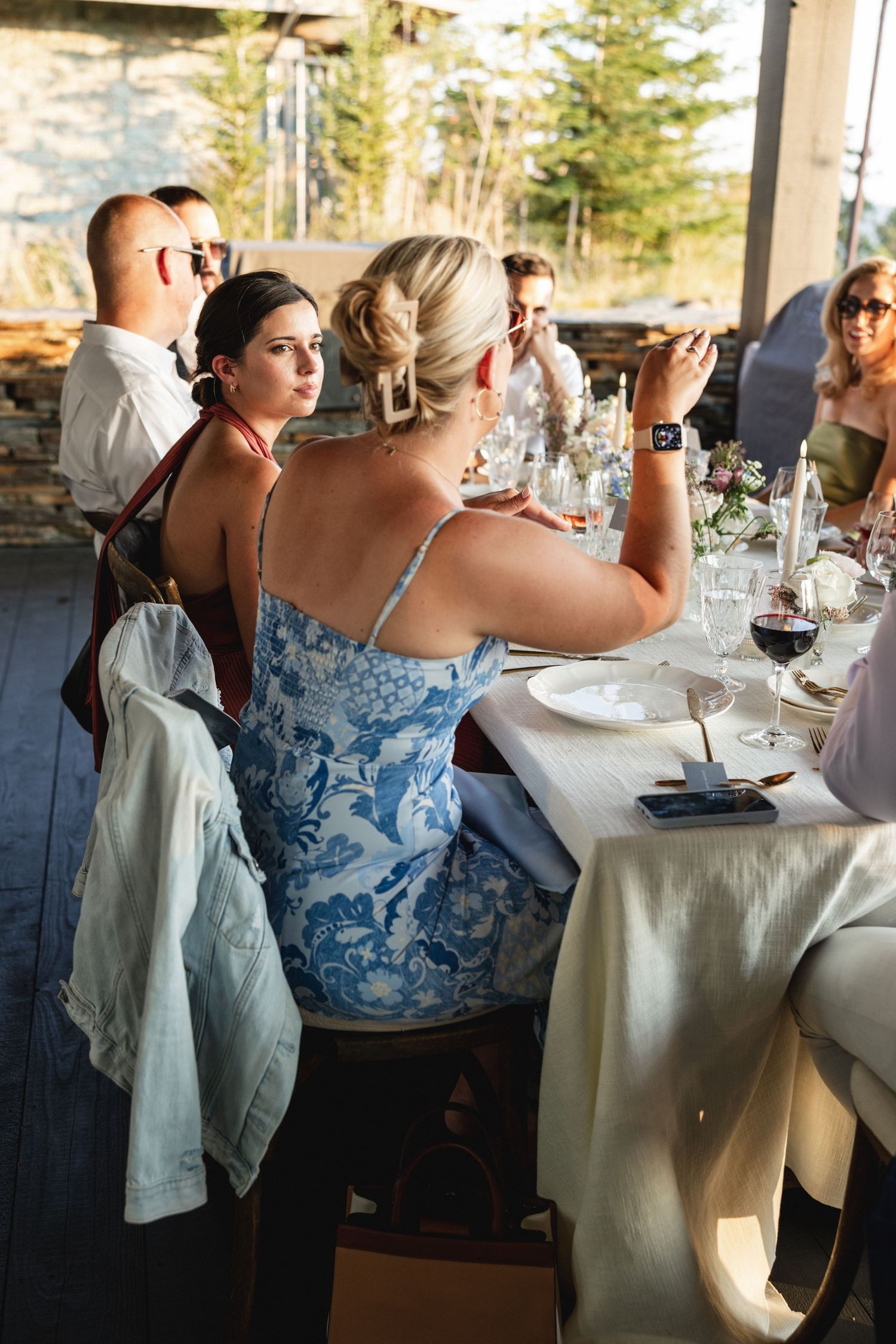 Guests seated at an elegant outdoor dinner party table, woman in blue floral dress taking a photo.