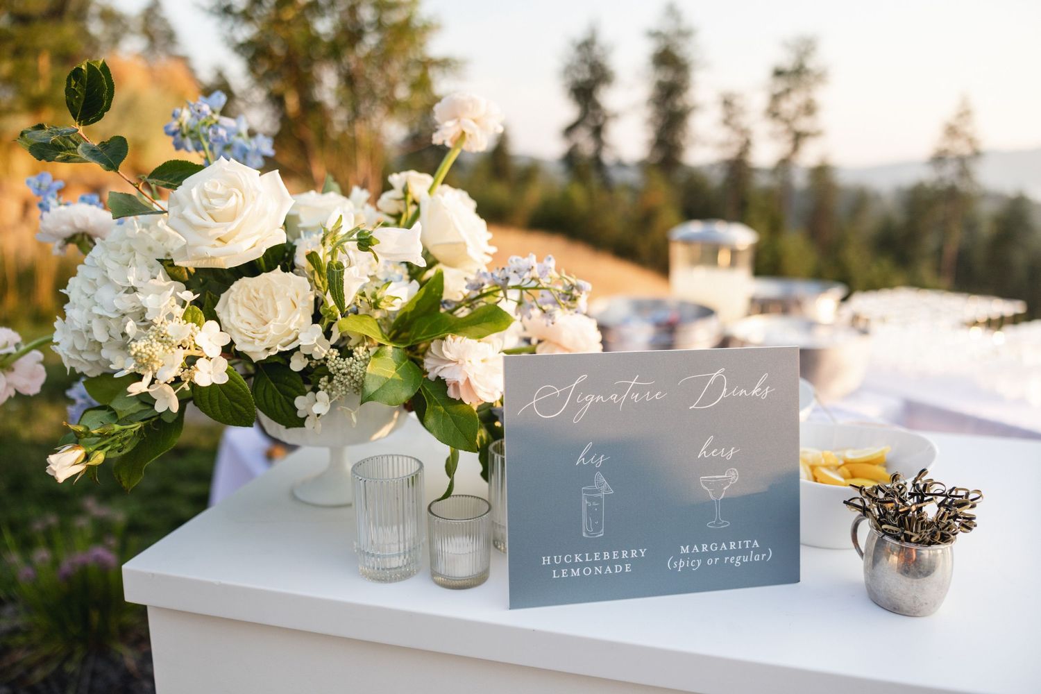 Wedding signature drinks sign beside white floral centerpiece with hydrangeas and roses.