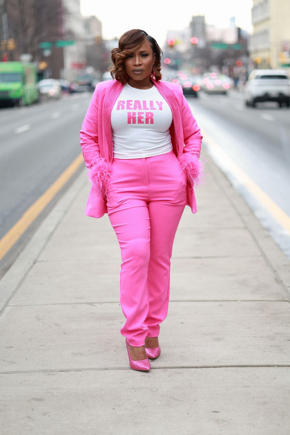 Woman in pink suit and heels walks confidently on city sidewalk wearing a white 'Really Her' graphic tee.
