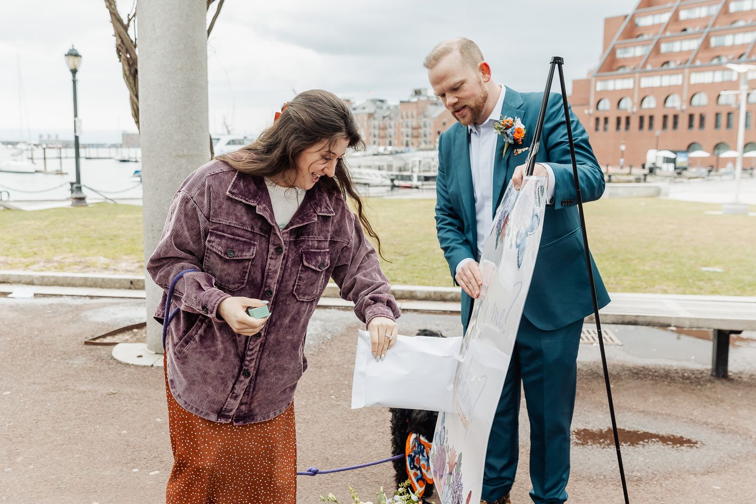 Couple opening wedding gifts outdoors near waterfront, woman in purple jacket and man in teal suit.