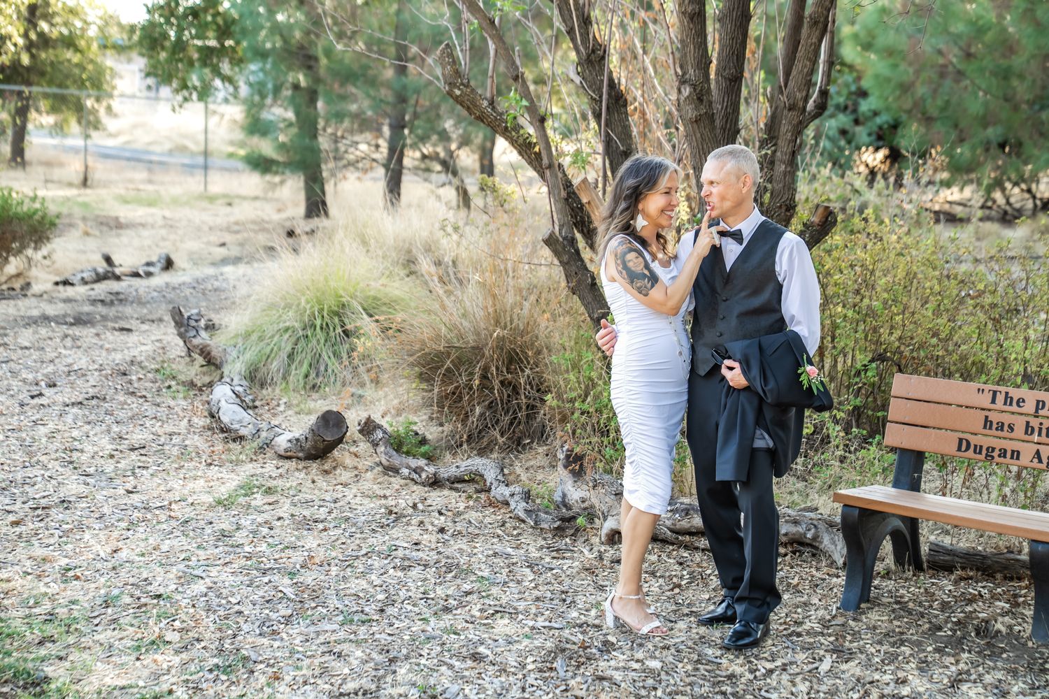 Couple in formal attire embracing outdoors near trees and a wooden bench on a gravel path.