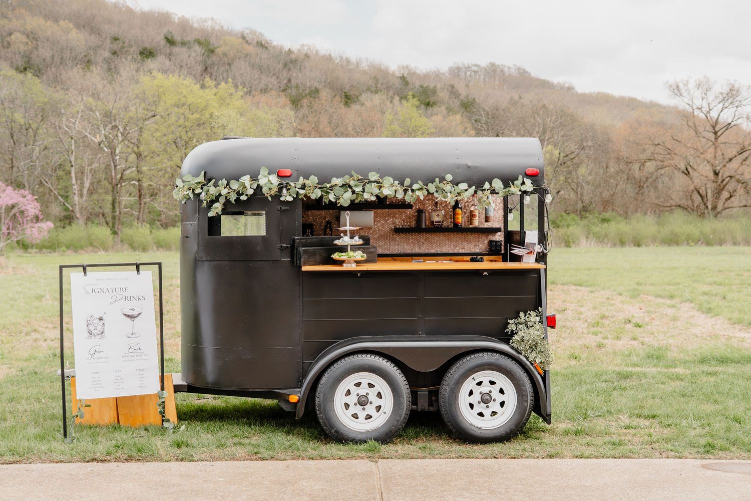 Black converted horse trailer bar with floral garland, serving as an outdoor wedding bar in a scenic green field.