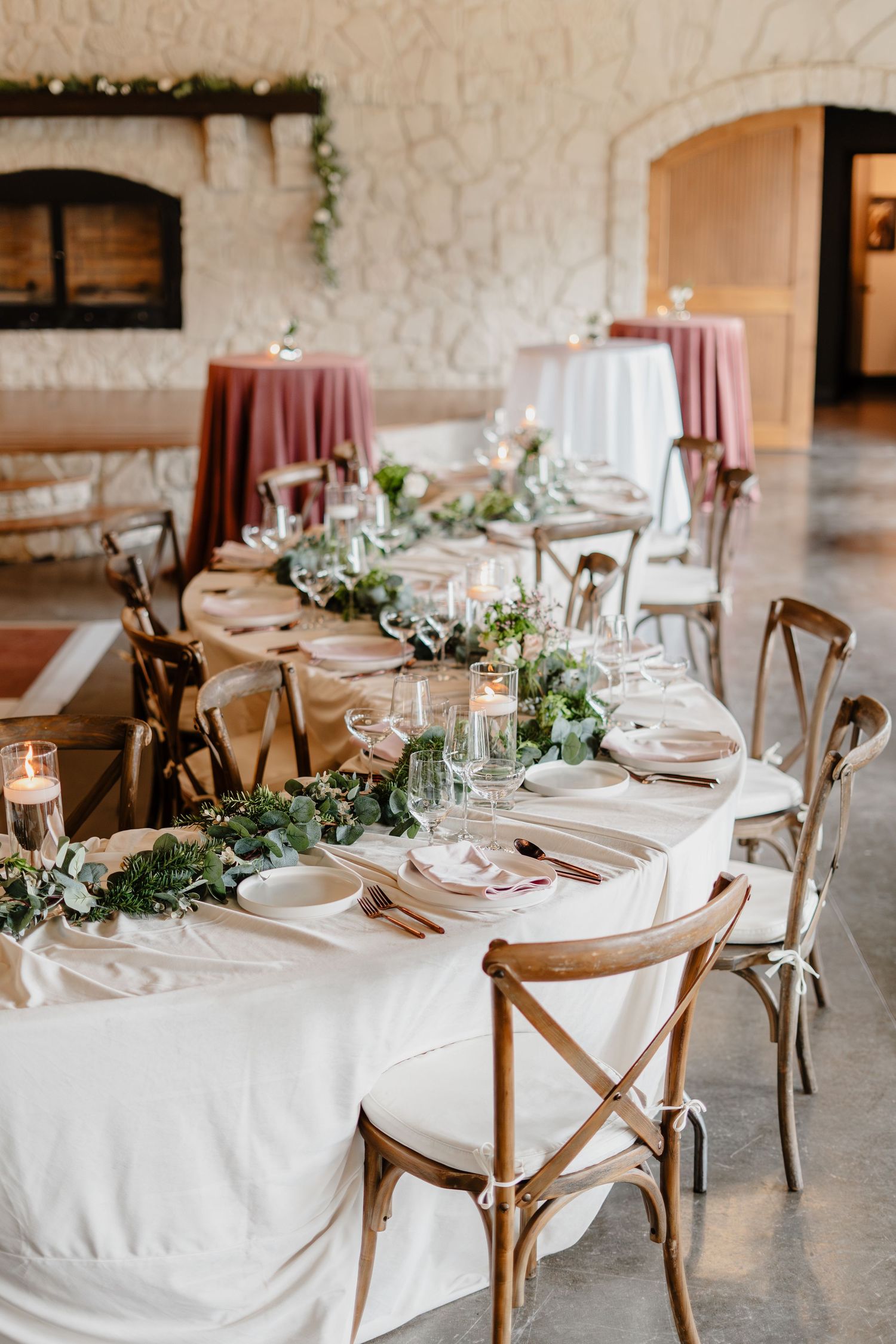 Elegant wedding reception table with white linen, greenery runner, candles, and rustic cross-back chairs in a stone venue.