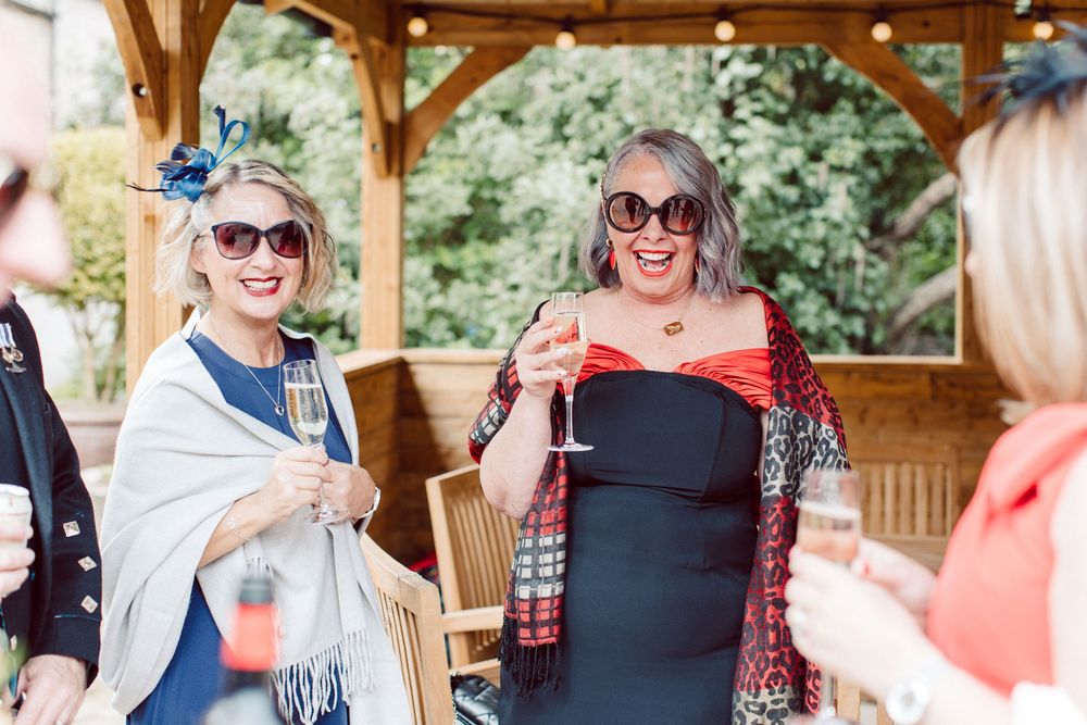 Wedding guests enjoying drinks and conversation at an outdoor reception under a wooden gazebo.