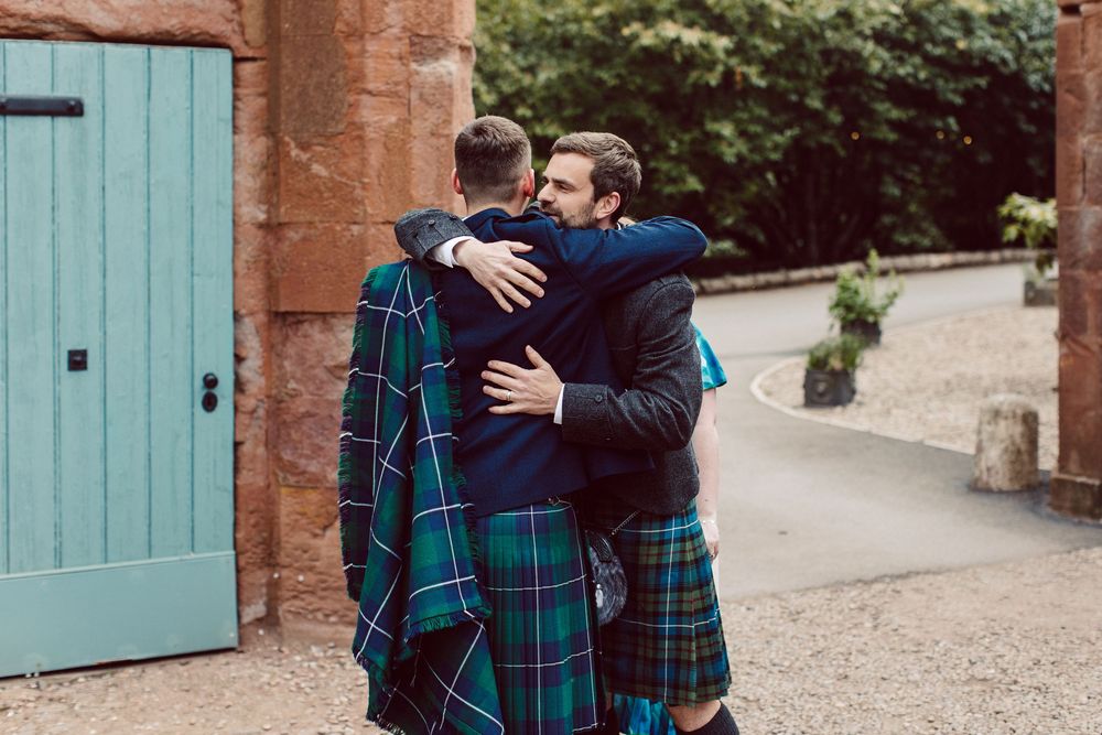 Two people wearing navy and plaid outfits embrace on a stone path with turquoise doors in the background.