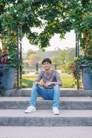 A person in casual attire sits on concrete steps framed by climbing vines and greenery in a garden setting.