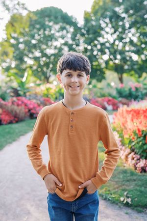 Young person in orange sweater and jeans poses outdoors near colorful autumn flowers in garden setting.