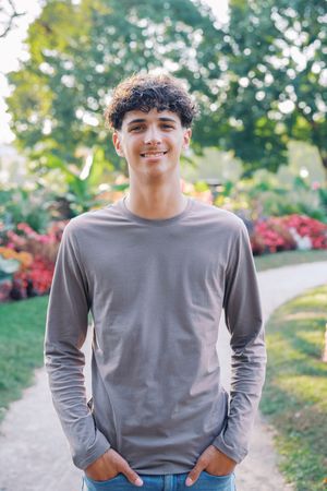 A person wearing a gray long sleeve shirt smiles while standing on a path with autumn foliage in the background.
