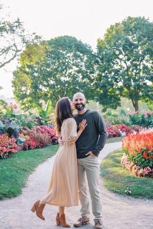 Couple embracing on garden path surrounded by vibrant pink and orange flowers on sunny day.