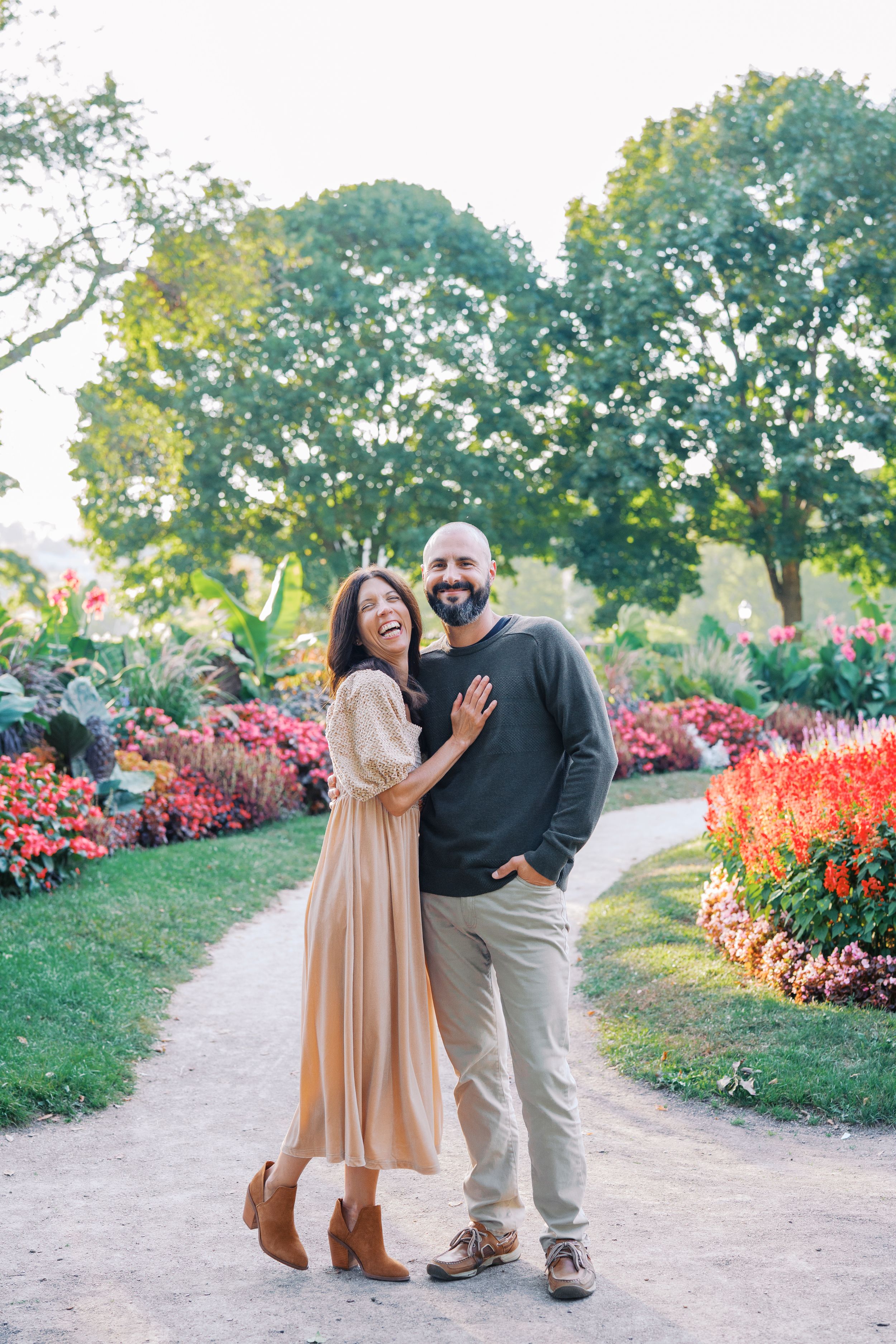 A couple poses together on a garden path with vibrant red flowers and greenery in the background.