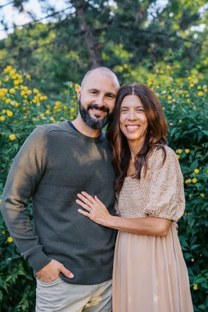 Two people embrace and laugh together outdoors with yellow flowers and trees in the background.
