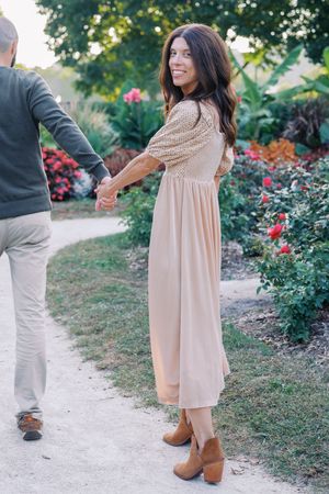 Woman in flowing beige dress walks along garden path with blooming flowers.