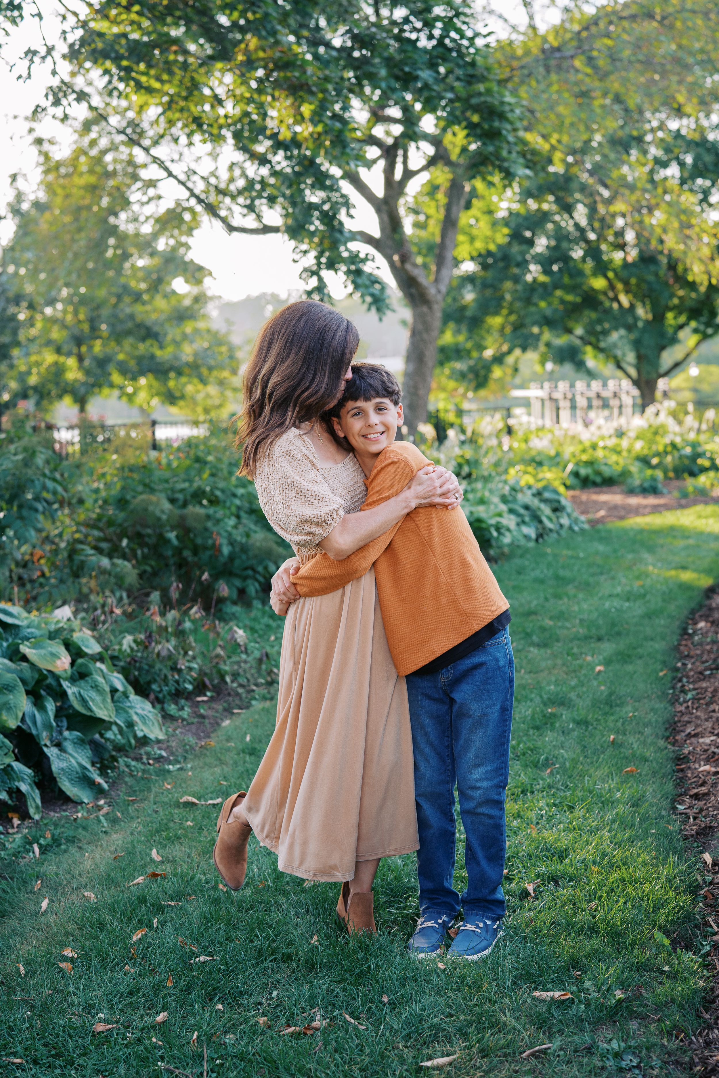 Three people share warm embraces while standing on a grassy path with trees and foliage in the background.