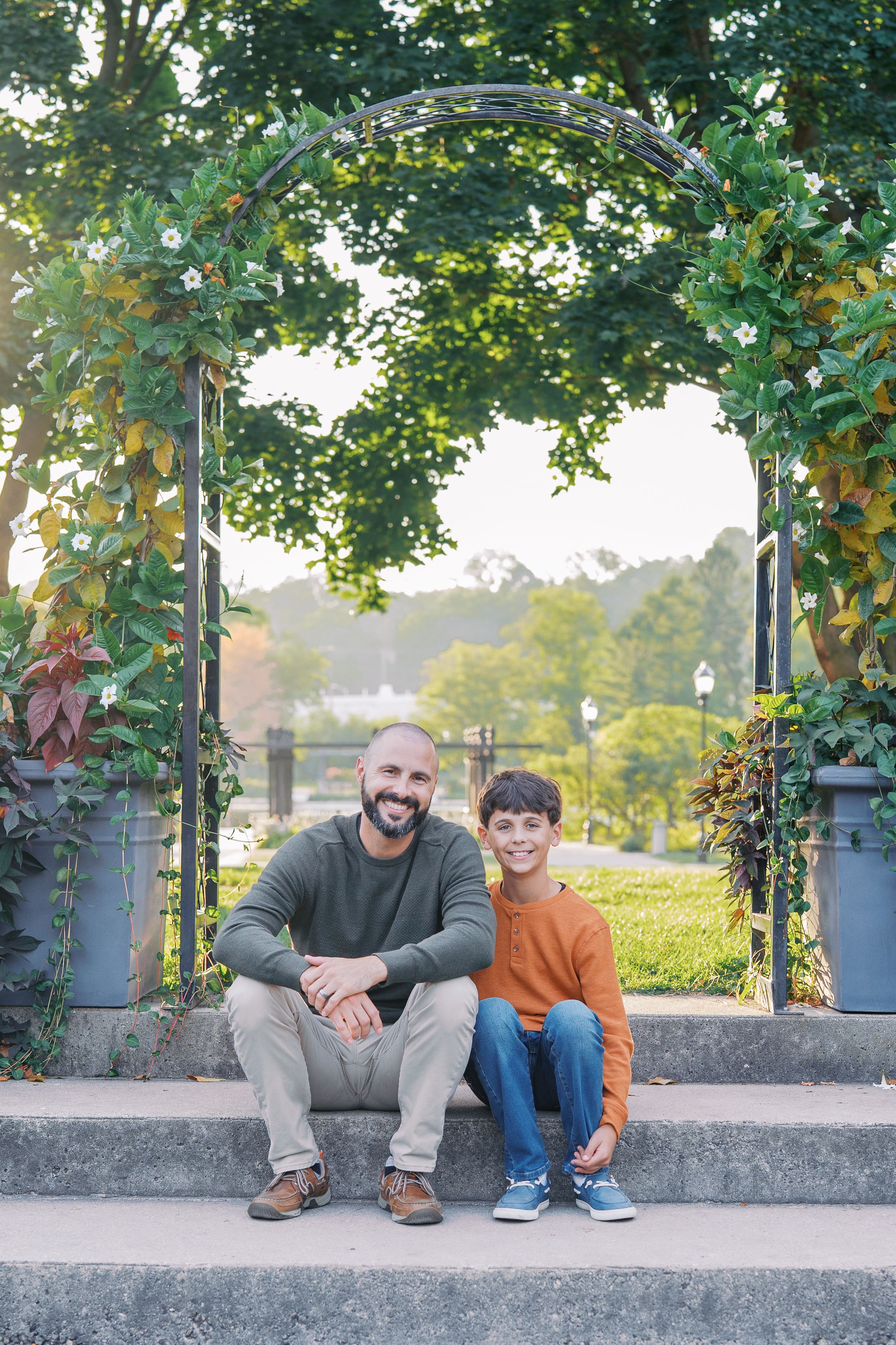 Two people sit together on stone steps under a decorative garden arch covered in green vines and leaves.