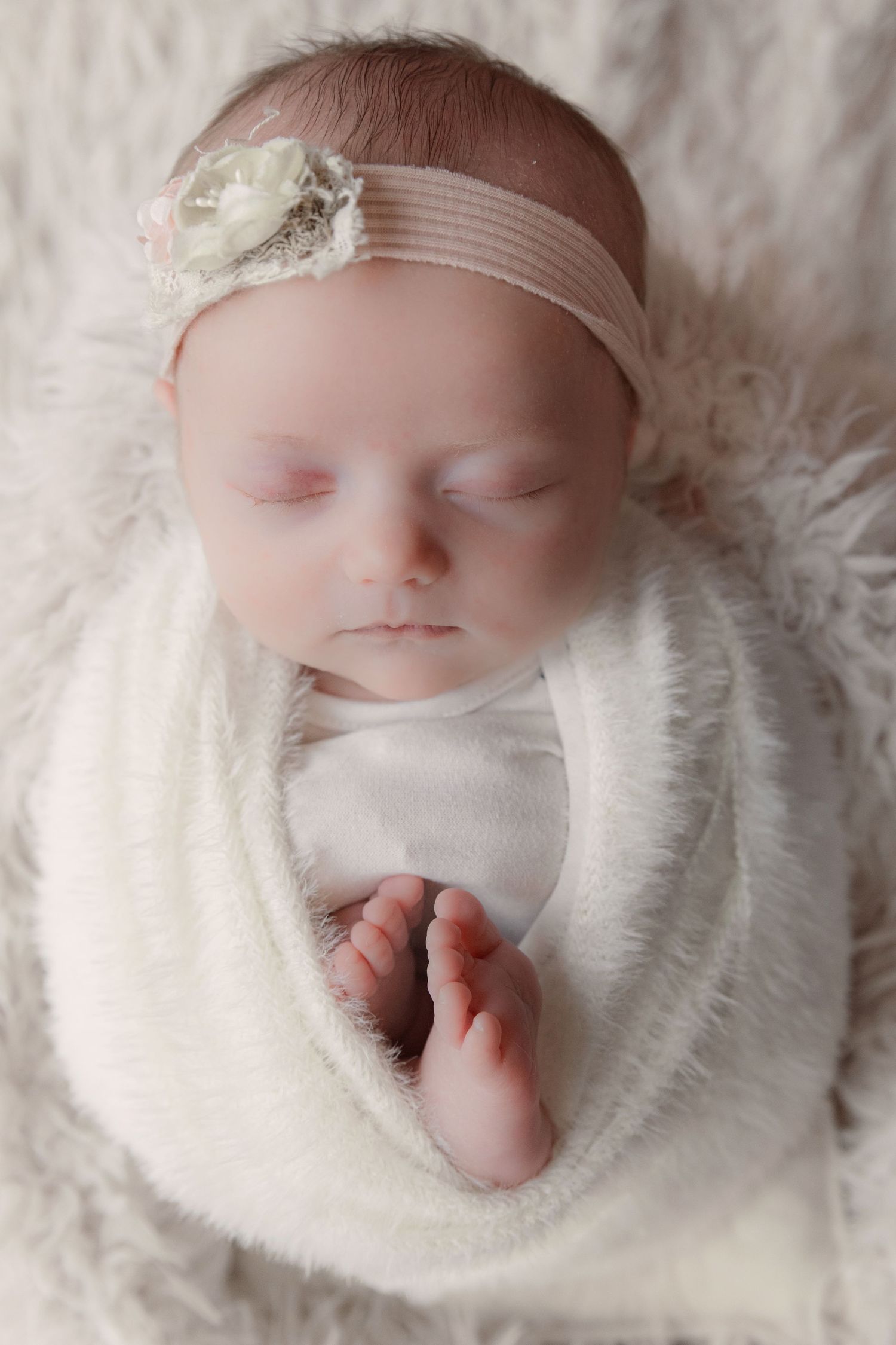 A sleeping newborn baby swaddled in white fabric lies on soft white fur blanket in a series of peaceful poses.