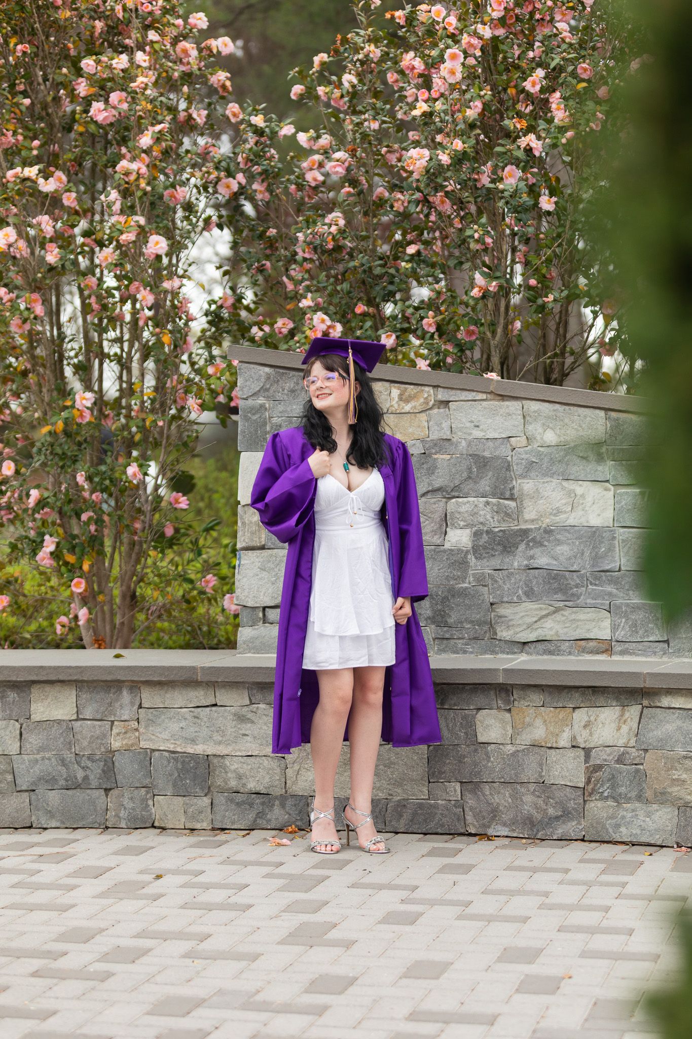 Young woman in purple graduation cap and gown poses outdoors by stone wall with blooming pink flowers.