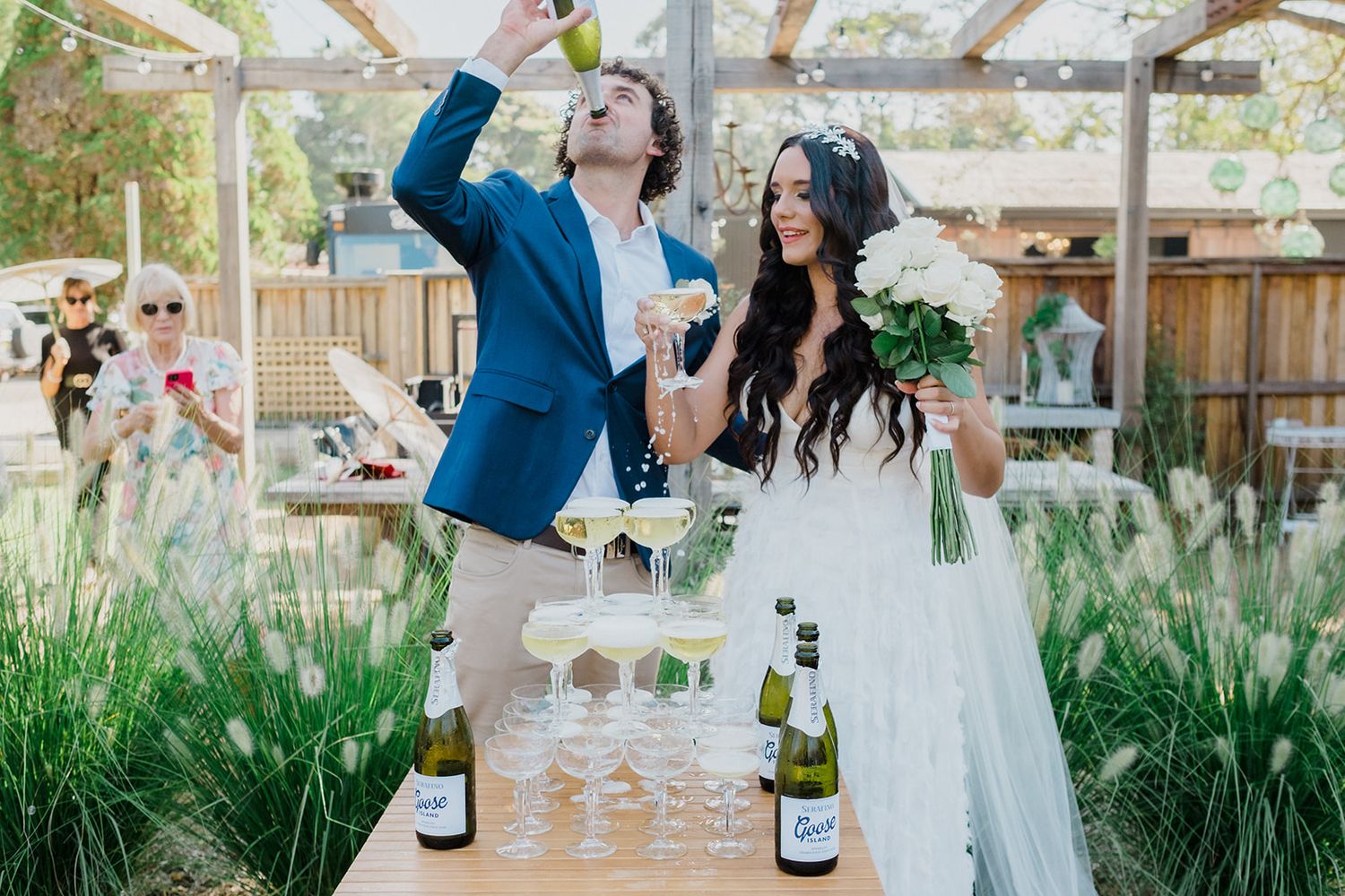 A couple celebrates at their outdoor garden wedding reception while pouring champagne at a rustic wooden table.
