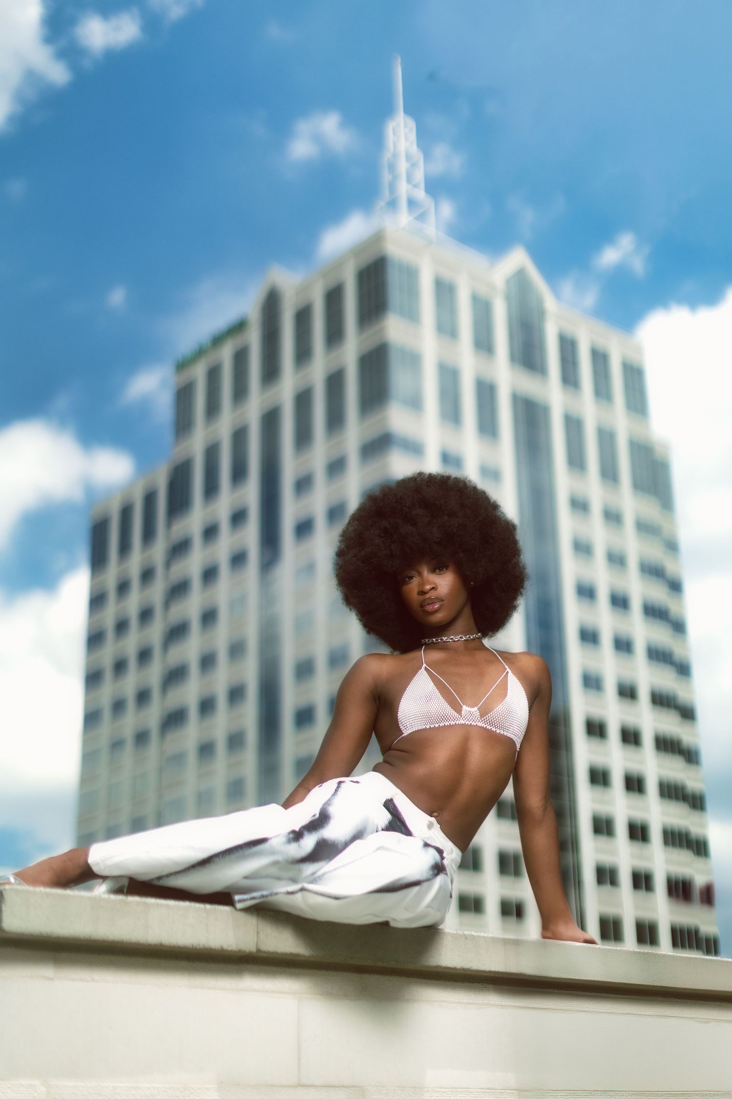 Stylish model in pink bikini and white pants poses against modern high-rise building on sunny day.