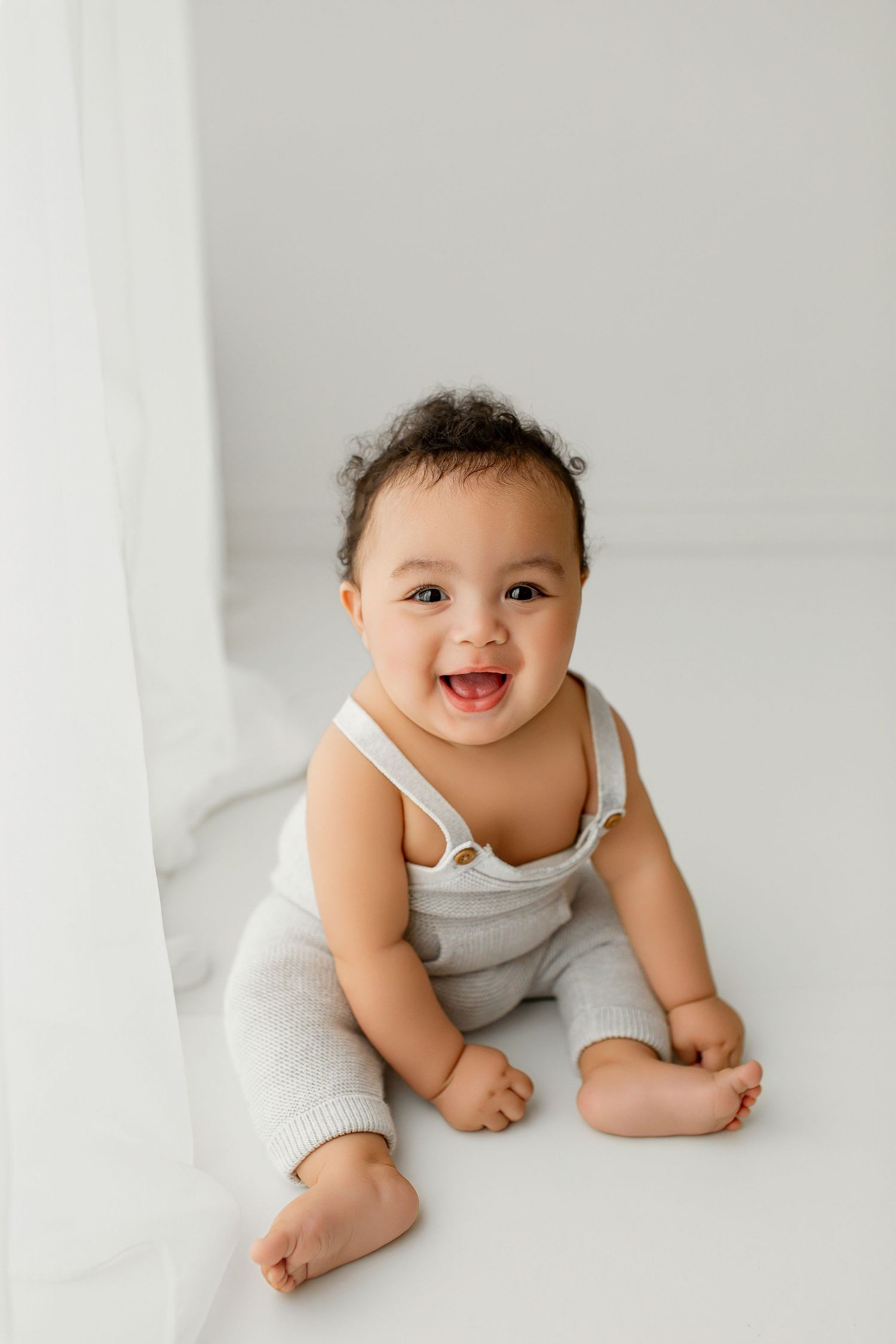 Laughing baby boy in light gray knit overalls sitting in white studio during sitter portrait session at Summerville SC