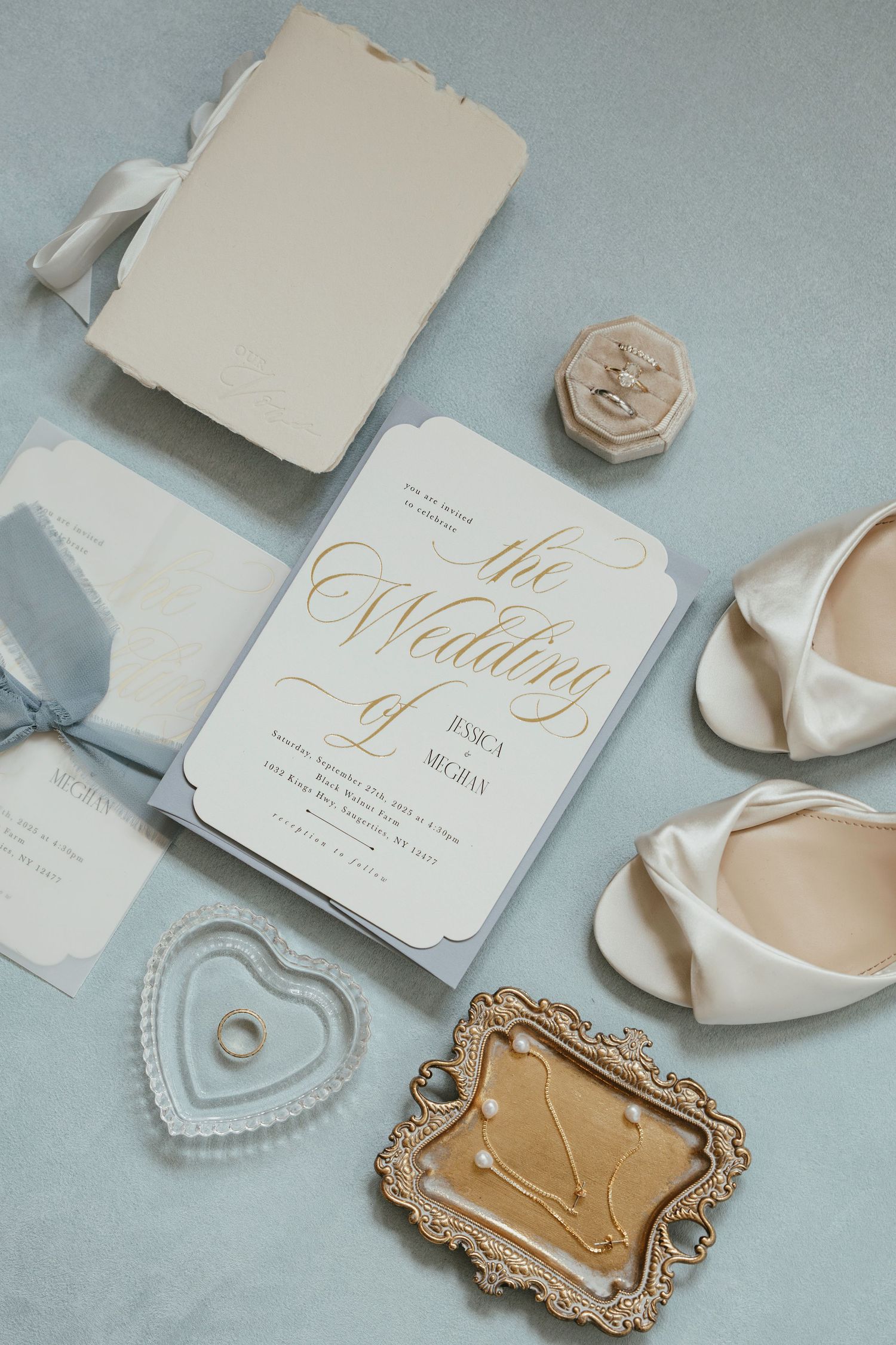 Elegant wedding details including white and gold invitation, cream heels, heart-shaped ring dish and vintage tray on blue surface.