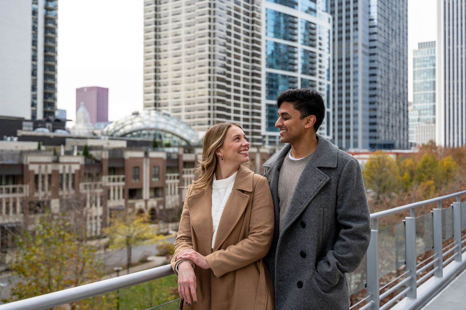 Two people in winter coats share a happy moment while walking along an urban railing with city skyscrapers in background.