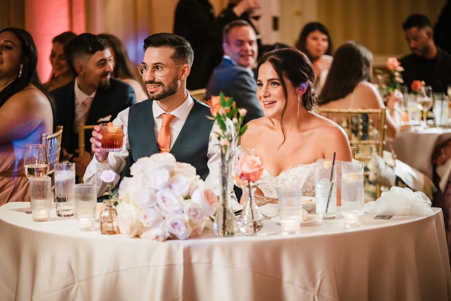Bride and groom smiling and listening to speeches at their sweetheart table during a Lenox Hotel Boston wedding reception