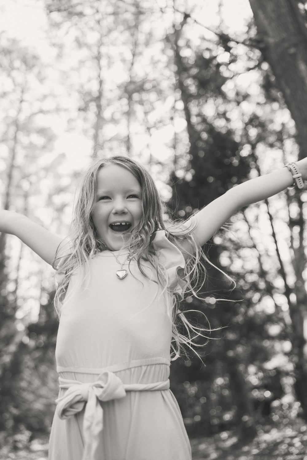 Joyful young girl with long hair laughing outdoors in forest, arms outstretched, wearing dress with bow, black and white photo.