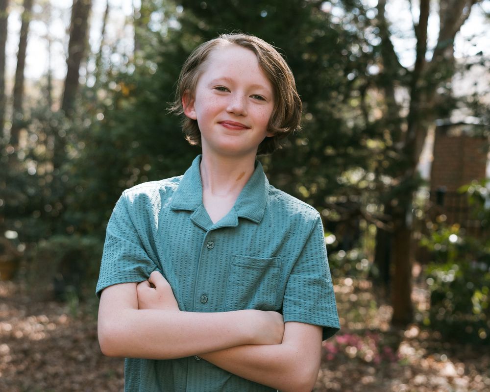 Smiling teen boy with crossed arms standing outdoors in a green button-up shirt surrounded by trees.