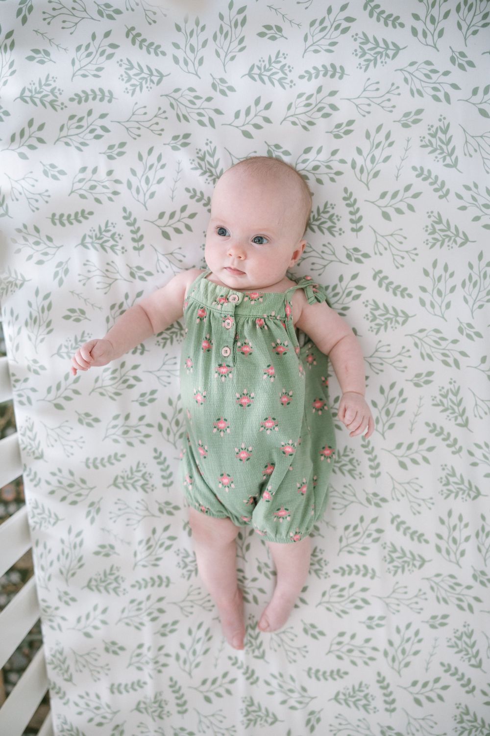 Baby girl in green floral romper lying in white leaf-print crib.
