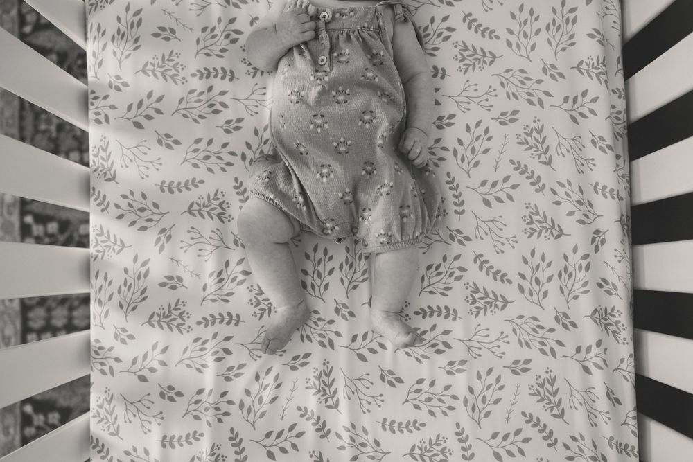 Black and white photo of a sleeping newborn baby lying in a white crib with leaf-patterned sheets.