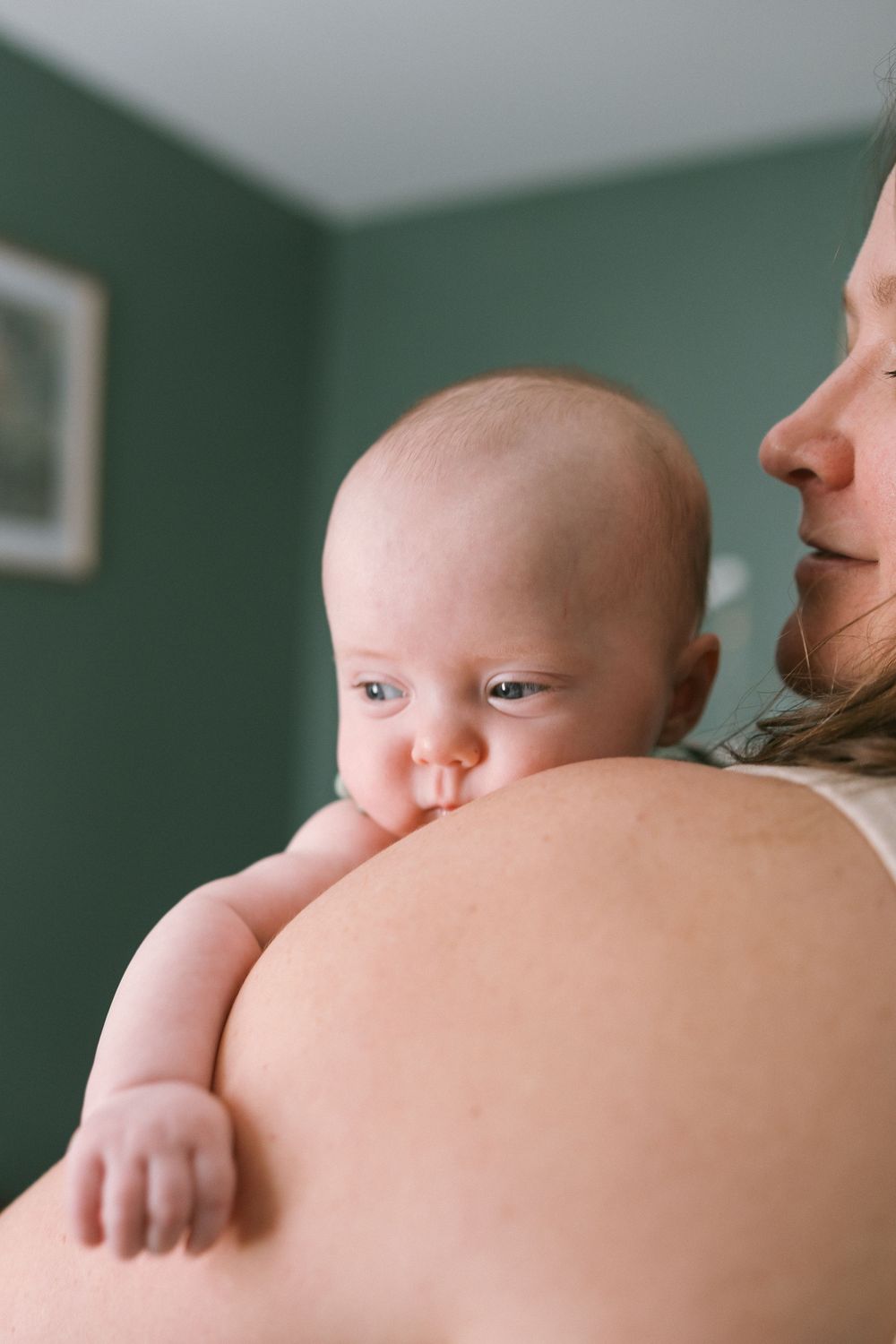 Mother tenderly holding newborn baby against her shoulder in room with sage green walls.