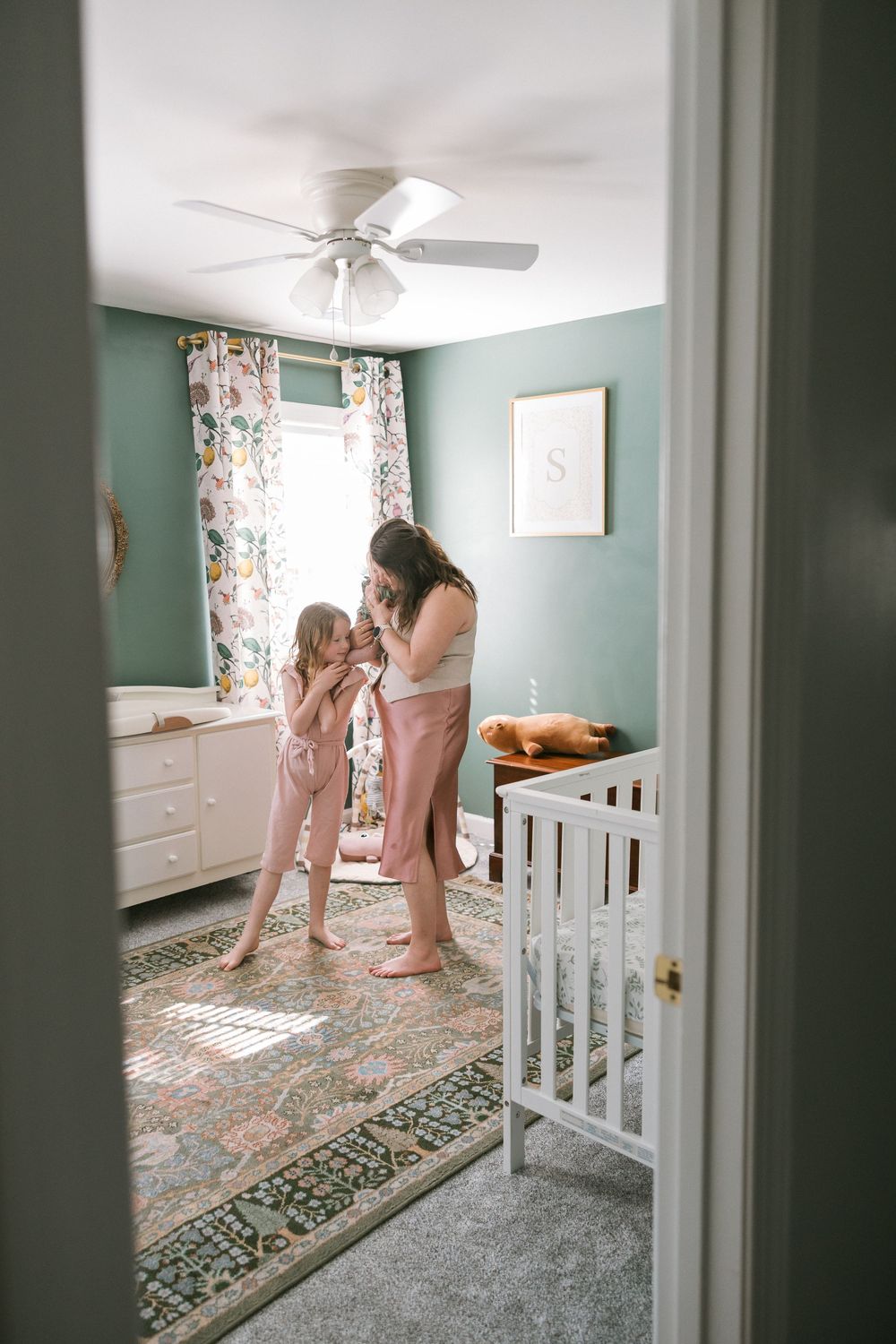 Mother and young daughter sharing a tender moment in a green nursery room with white crib and floral curtains.