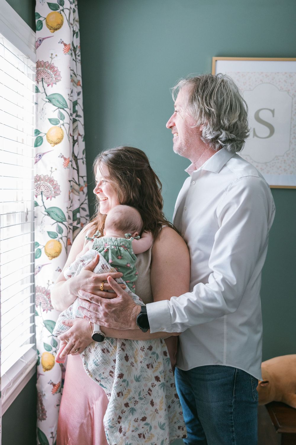 Happy couple holding newborn baby by window with floral curtains in nursery with green walls.