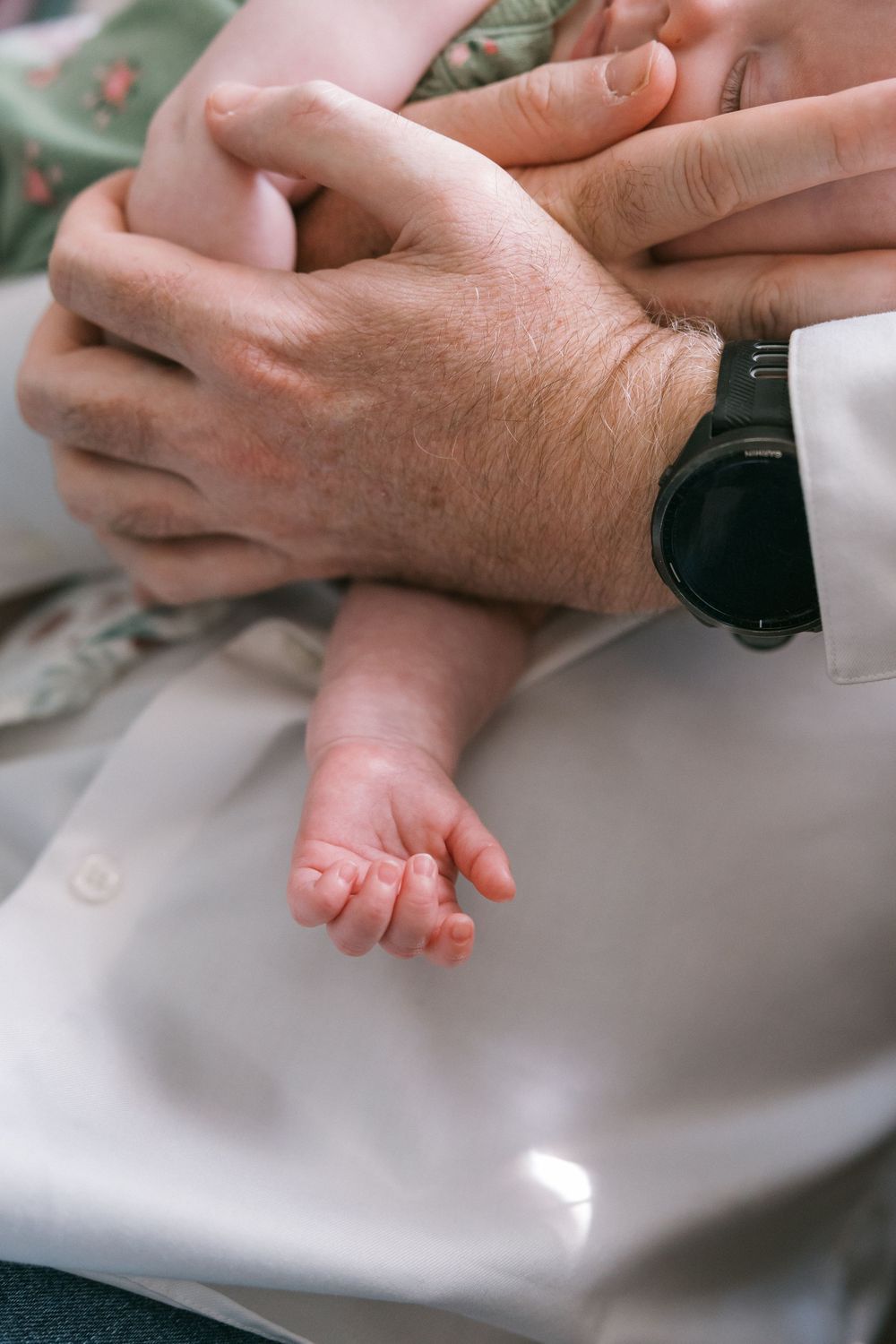 Close-up of a newborn baby's tiny hand and foot resting on an adult's lap, with a man's hand wearing a watch nearby.