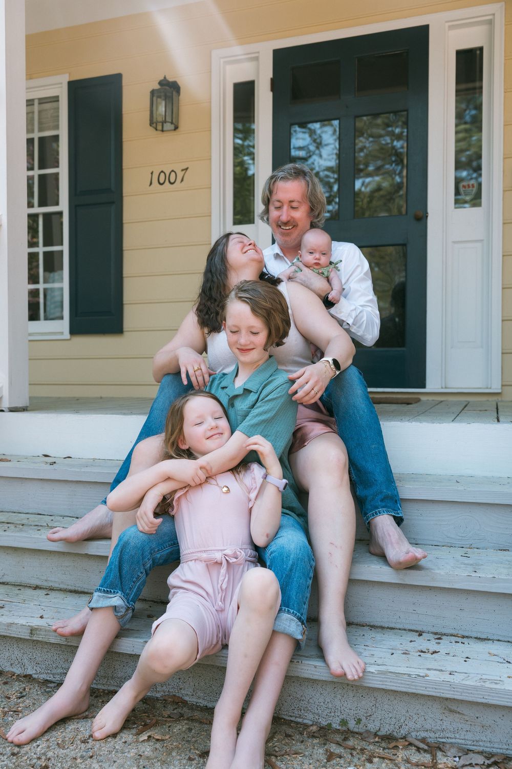 Happy family of five sitting on front porch steps of yellow house numbered 1007, parents with three children including newborn.