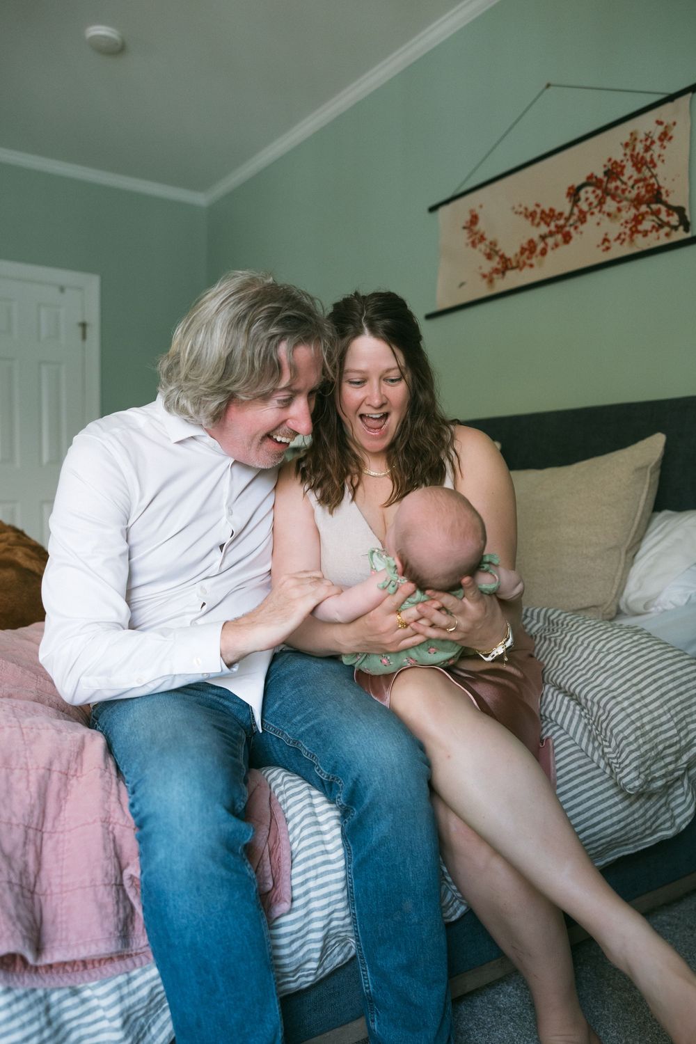 Happy couple sitting on bed laughing while holding their newborn baby in a cozy bedroom.