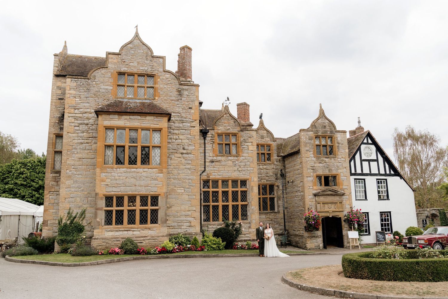 A grand, historic stone manor house with Tudor-style architectural features, surrounded by landscaped gardens.