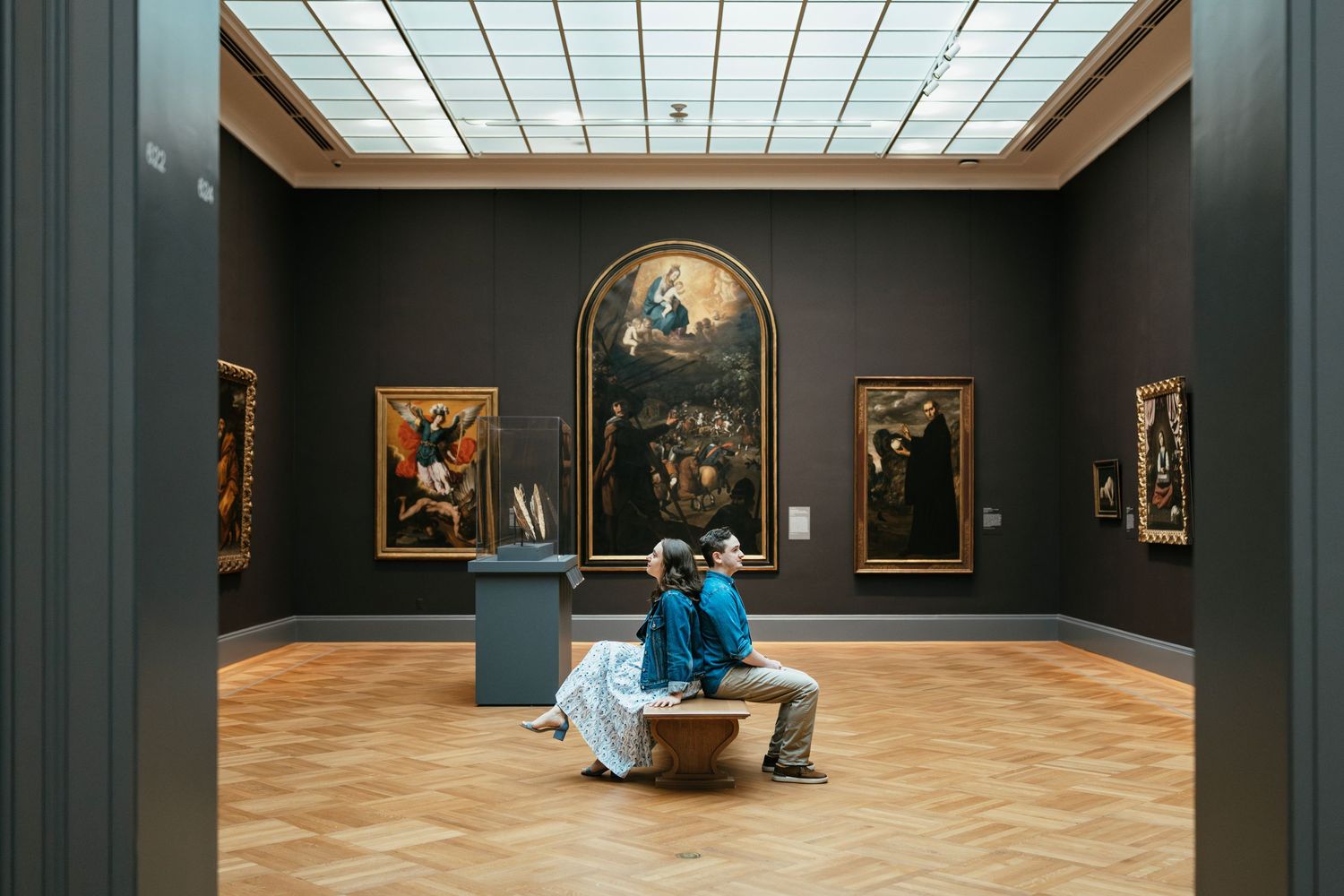 Museum visitors sit on bench viewing classical paintings in dark green gallery with skylight at Metropolitan Museum of Art.