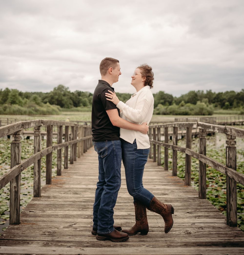 Couple embracing on a wooden bridge boardwalk surrounded by lush greenery under a cloudy sky.