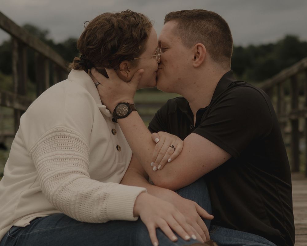 Couple sharing a romantic kiss outdoors on a bridge, woman in white sweater and man in black shirt.