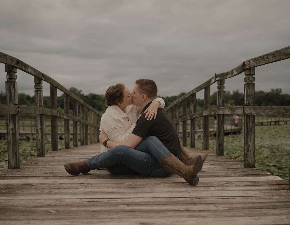 Couple sitting on a wooden bridge dock kissing romantically with cloudy skies and water in background.