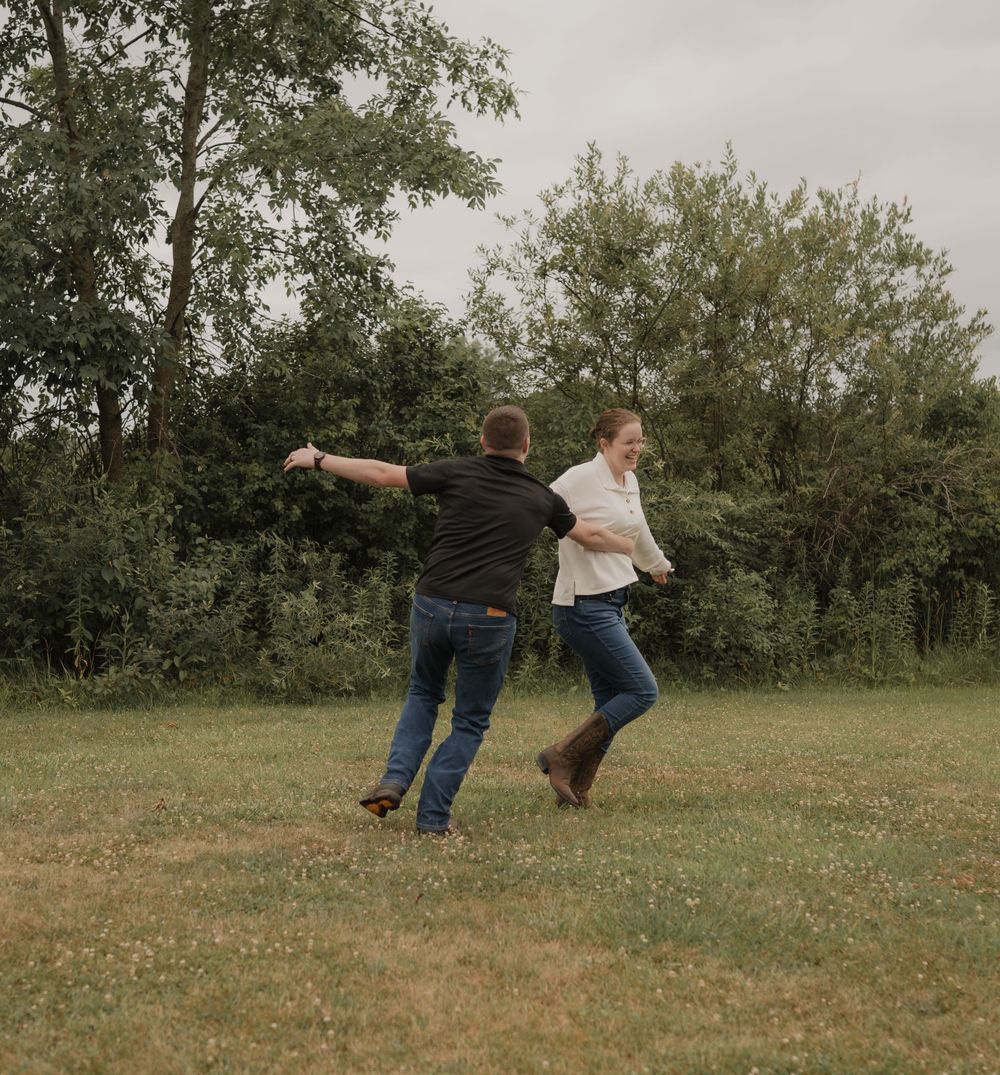 Couple dancing and spinning together on green grass field with trees in background on overcast day.