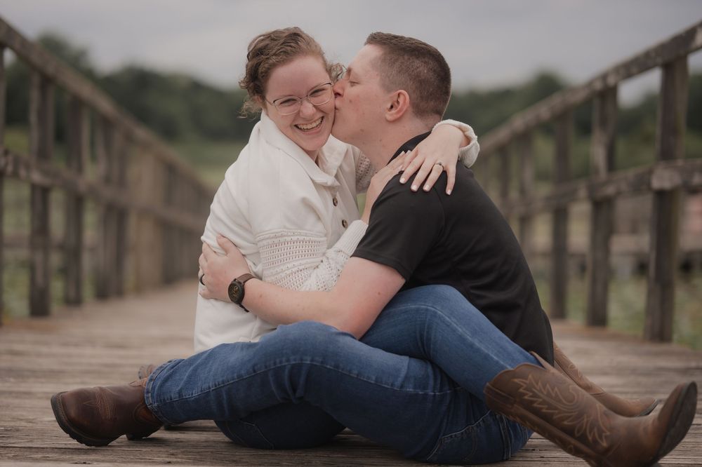 Couple sitting on wooden bridge, man kissing woman's cheek, both smiling and wearing jeans outdoors.