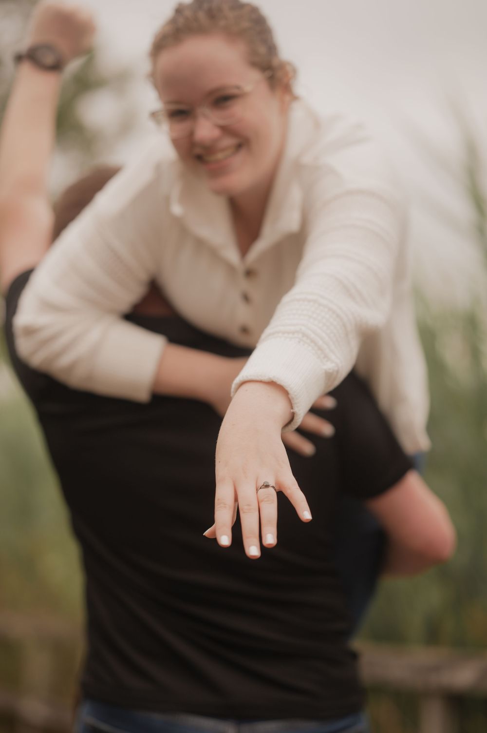 Smiling woman being lifted by partner showing off engagement ring outdoors.