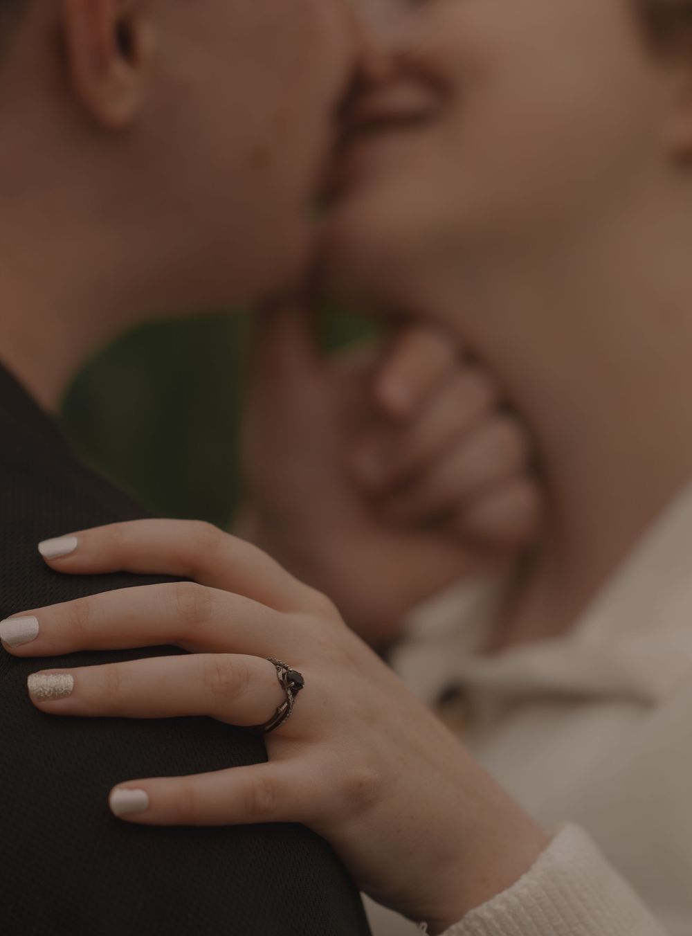 Close-up of womans hand with black engagement ring resting on partners shoulder while kissing.