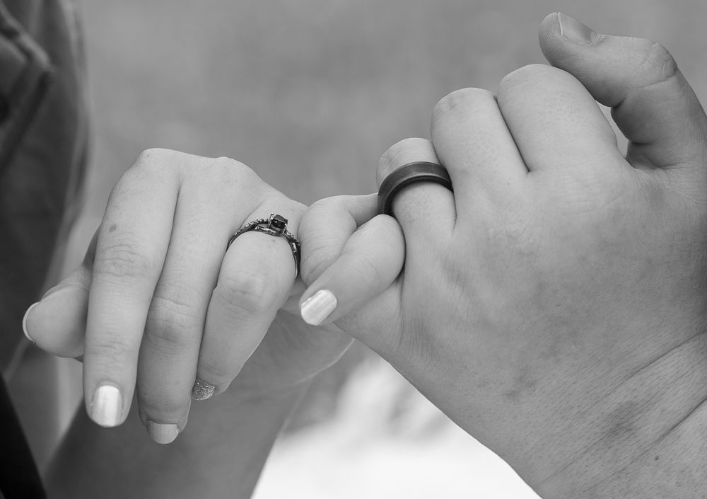 Black and white photo of couple holding pinkies, woman wearing diamond engagement ring, man wearing black wedding band.