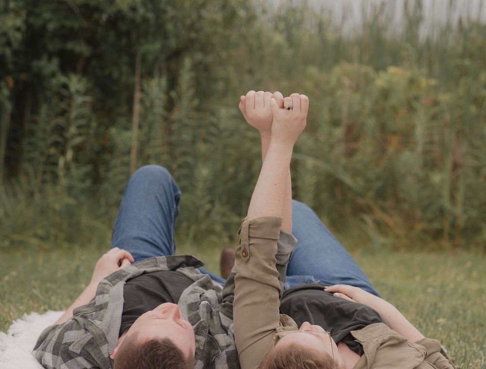 Couple lying on grass holding hands raised up, wearing jeans and casual jackets, with green trees in background.