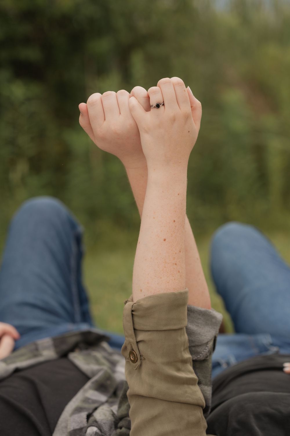 Two people holding hands raised up while lying in grass outdoors, one wearing a ring.