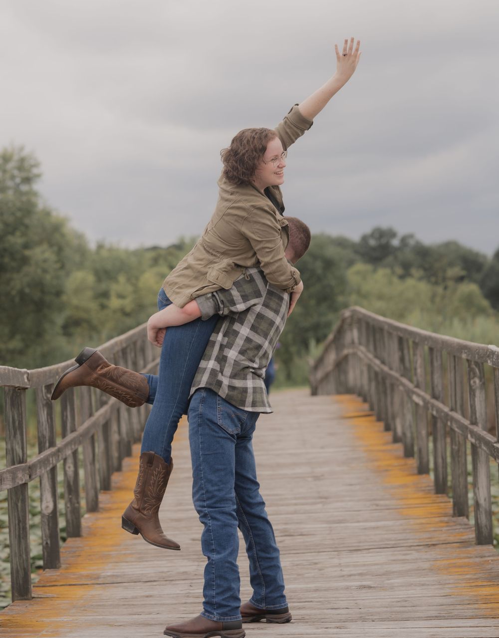 A man lifts a woman on a wooden bridge, she reaches up joyfully, both wearing jeans and boots outdoors.